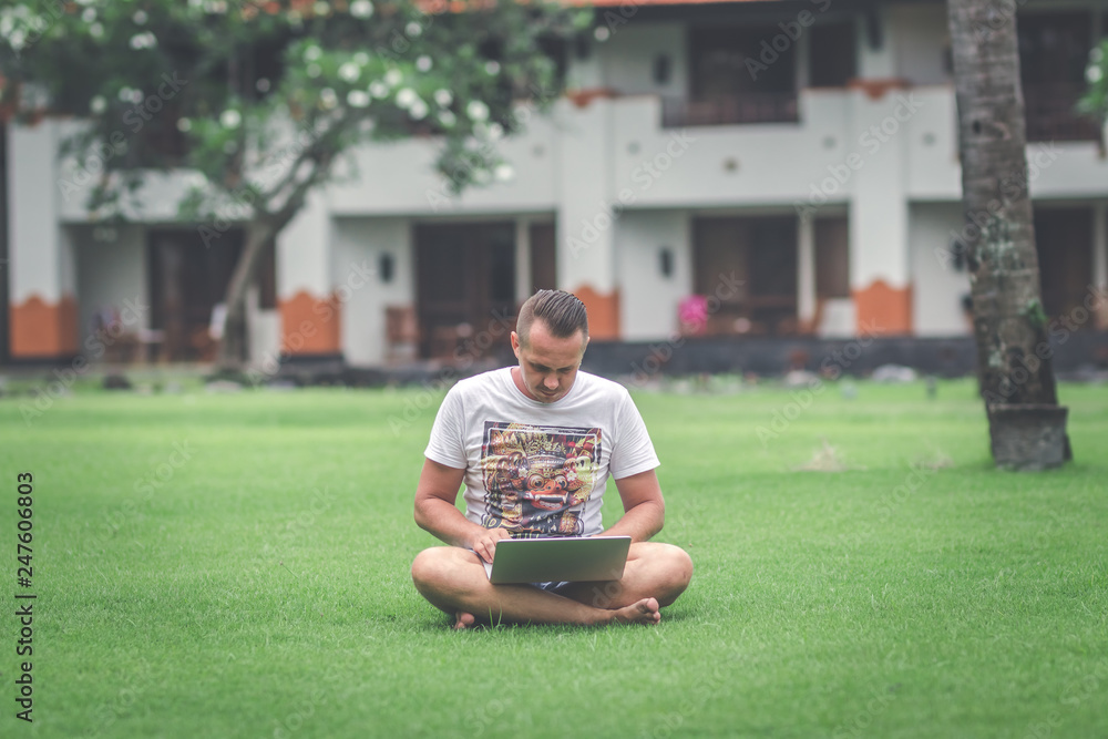 Fototapeta premium Young man in the green tropical park working online with laptop. Freelancer man on Bali island concept.
