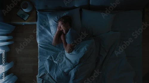 Top View of Handsome Young Man Sleeping Cozily on a Bed in His Bedroom at Nigh, He Tosses and Turns. Blue Nightly Colors with Cold Weak Lamppost Light Shining Through the Window. photo