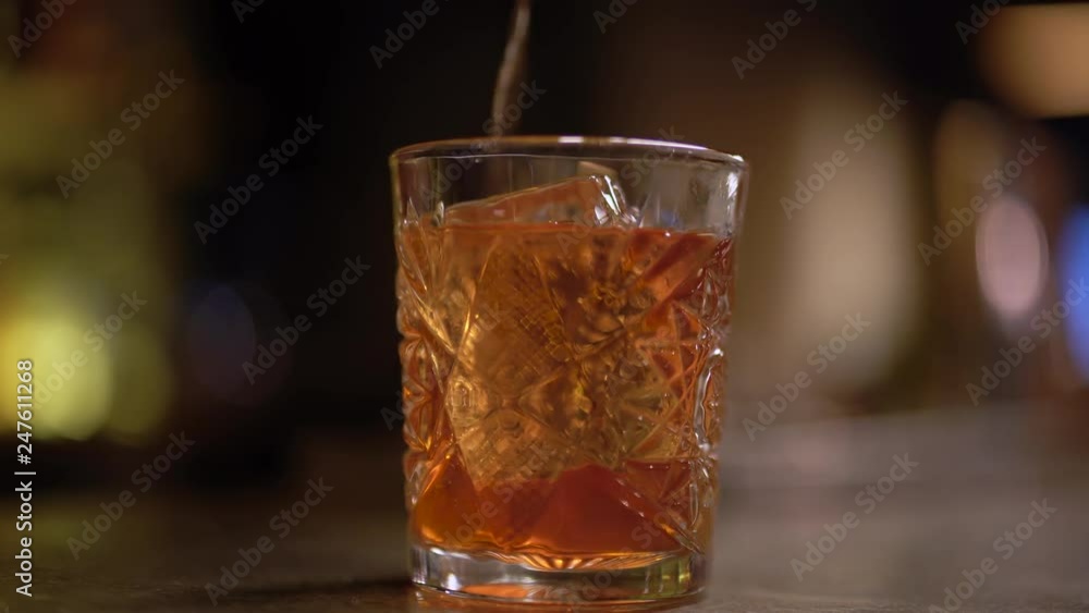 Alcohol shot with one piece of ice at the table close up. Orange liquid pouring in the glass slowly Alcohol shot with one piece of ice at the table close up. Orange liquid pouring in the glass slowly