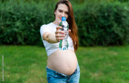 Crop of stylish pregnant woman in jeans and white shirt showing bootle of water in hand and showing at camera. Brunette mom posing in park, spending time outdoors. Concept of health pregnant.