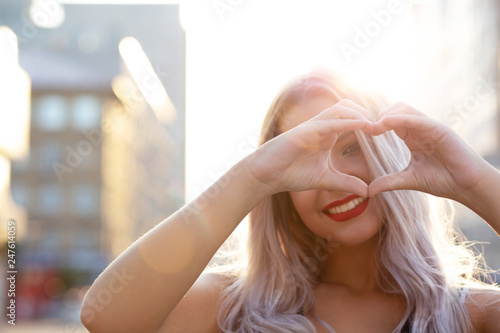 Joyful blonde model with red lips making heart shape with her fingers at the street. Empty space