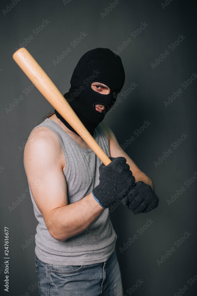 Bandit, the criminal in a black mask and in a gray T-shirt with a painless bat. Robber with a weapon. A man in a balaclava and a gray mask on a gray background Bandit, the criminal in a black mask and in a gray T-shirt with a painless bat. Robber with a weapon. A man in a balaclava and a gray mask on a gray background
