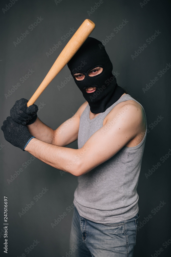 Bandit, the criminal in a black mask and in a gray T-shirt with a painless bat. Robber with a weapon. A man in a balaclava and a gray mask on a gray background Bandit, the criminal in a black mask and in a gray T-shirt with a painless bat. Robber with a weapon. A man in a balaclava and a gray mask on a gray background