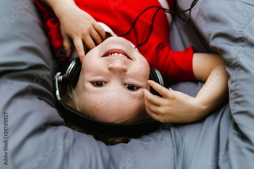 Cute little smart Caucasian girl in casual red dress and headphones listening to music while lying on the chair in the room. The girl lies upside down and laughing