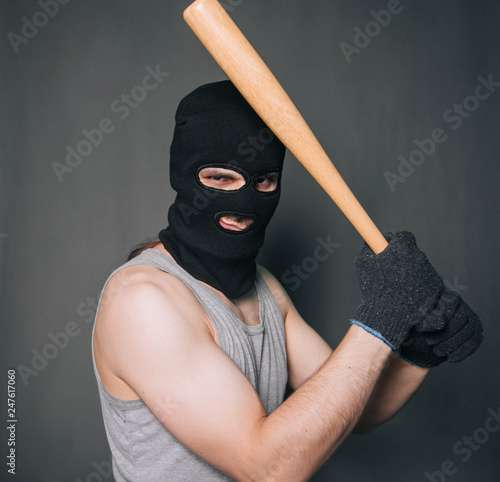 Bandit, the criminal in a black mask and in a gray T-shirt with a painless bat. Robber with a weapon. A man in a balaclava and a gray mask on a gray background photo