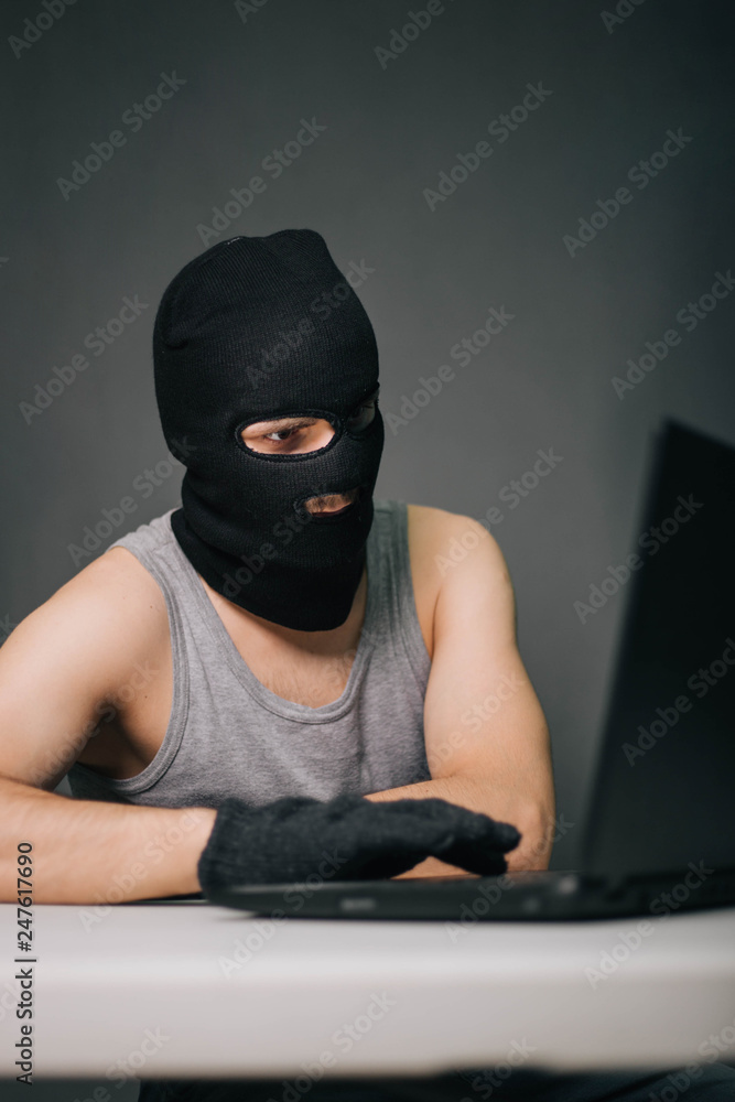 A thief in a black mask stole a laptop. A man in a balaclava and a computer in hand on a gray background. The hacker is hacking the computer. Steals information. A young man breaks the law. A thief in a black mask stole a laptop. A man in a balaclava and a computer in hand on a gray background. The hacker is hacking the computer. Steals information. A young man breaks the law.
