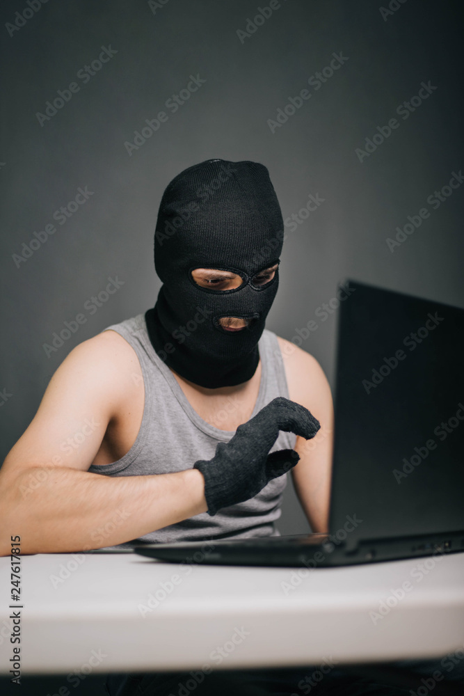 A thief in a black mask stole a laptop. A man in a balaclava and a computer in hand on a gray background. The hacker is hacking the computer. Steals information. A young man breaks the law. A thief in a black mask stole a laptop. A man in a balaclava and a computer in hand on a gray background. The hacker is hacking the computer. Steals information. A young man breaks the law.