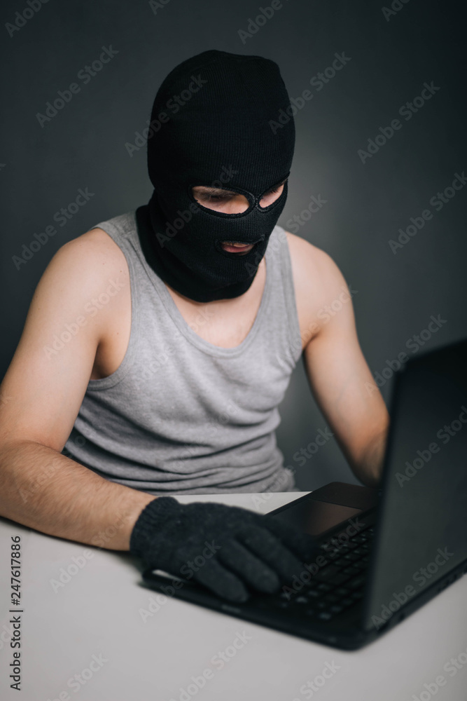 A thief in a black mask stole a laptop. A man in a balaclava and a computer in hand on a gray background. The hacker is hacking the computer. Steals information. A young man breaks the law. A thief in a black mask stole a laptop. A man in a balaclava and a computer in hand on a gray background. The hacker is hacking the computer. Steals information. A young man breaks the law.