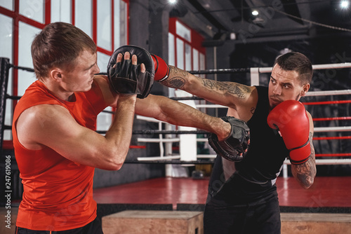 Counter punch. Strong tattooed athlete in sports clothing training on boxing paws with partner opposite boxing ring