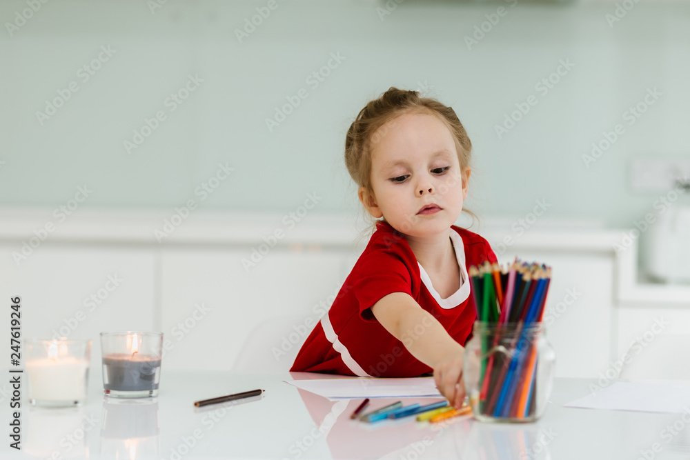 Cute little Caucasian girl in casual red dress draws with colored pencils in a bright room. Preparing a preschooler for school Cute little Caucasian girl in casual red dress draws with colored pencils in a bright room. Preparing a preschooler for school
