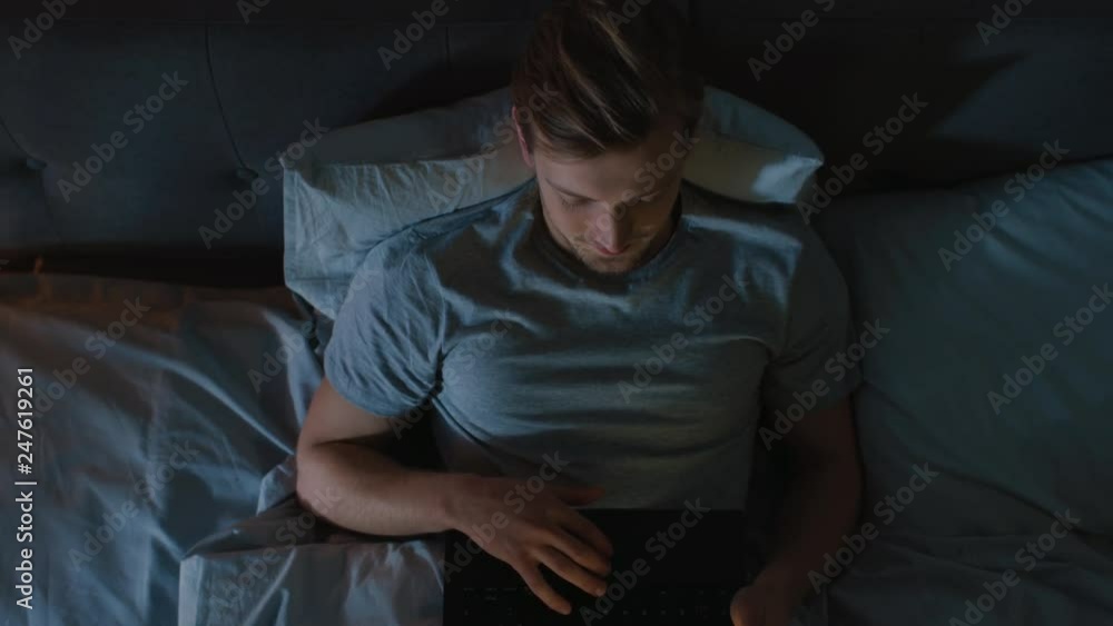 Top View Young Man in Bed Working on a Laptop Computer at Night. Student Getting Ready to Exams, Exceptionally Dedicated Project Manager Finishing Work in Bed at Night. Top View Young Man in Bed Working on a Laptop Computer at Night. Student Getting Ready to Exams, Exceptionally Dedicated Project Manager Finishing Work in Bed at Night.