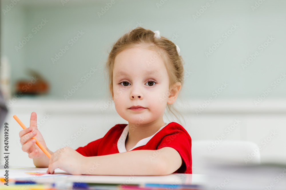 Cute little Caucasian girl in casual red dress draws with colored pencils in a bright room. Preparing a preschooler for school Cute little Caucasian girl in casual red dress draws with colored pencils in a bright room. Preparing a preschooler for school