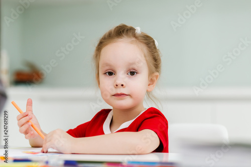 Cute little Caucasian girl in casual red dress draws with colored pencils in a bright room. Preparing a preschooler for school