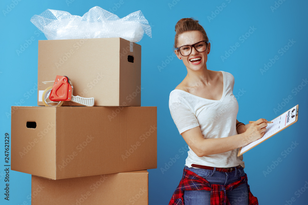 happy woman near cardboard box marking moving checklist on blue happy woman near cardboard box marking moving checklist on blue