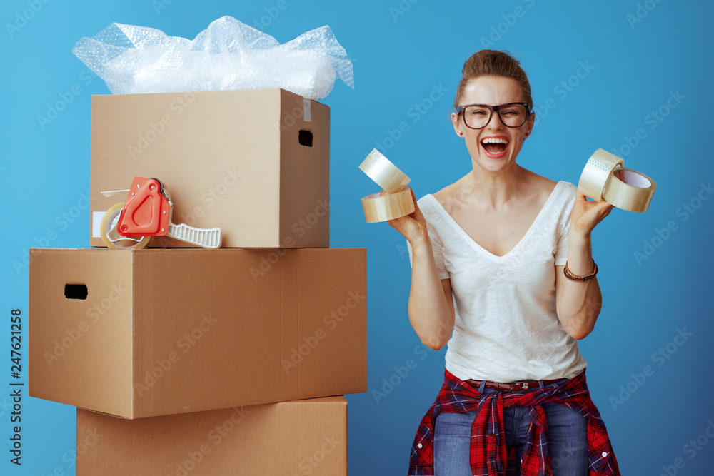 smiling woman near cardboard box showing adhesive tapes on blue - obrazy, fototapety, plakaty 