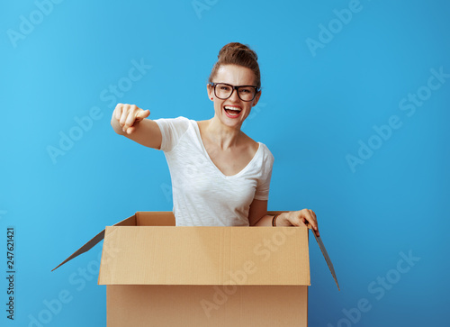 smiling young woman in cardboard box pointing in camera on blue