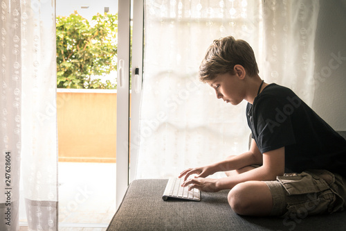 Beautiful blond child sitting on the couch at home using computer. Young boy relaxing with laptop free time online games. Teen working and chat with internet friends. Student doing homework on the web