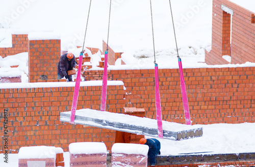 Reinforced concrete slab, crane transportation. Tape slings are used. Place of action - construction of a brick house. The visible construction workers in hard hats. Winter, lies snow.