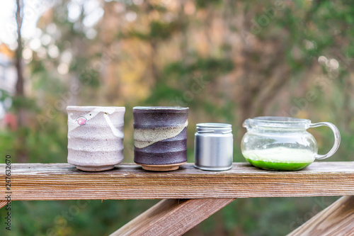 Glass teapot on wooden railing outside on fence of garden with two cups and Japanese vibrant green matcha tea color in morning closeup photo