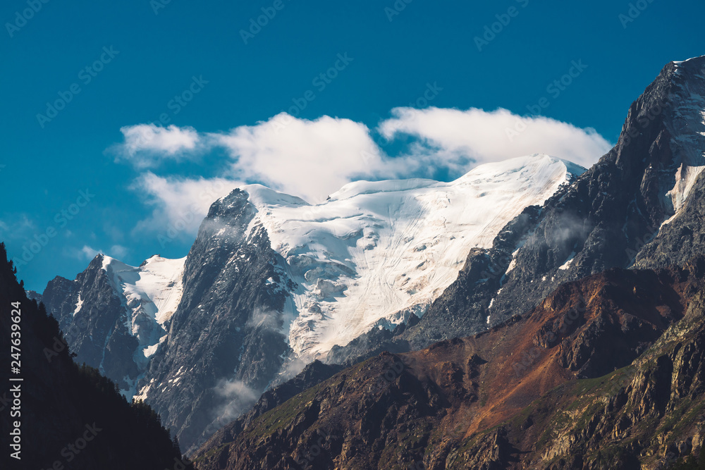 Low clouds and mist on top of mountain range. Glacier under clear blue sky. Snowy mountain peak in sunny day. Giant rocky ridge with snow. Atmospheric minimalist highland landscape of majestic nature.