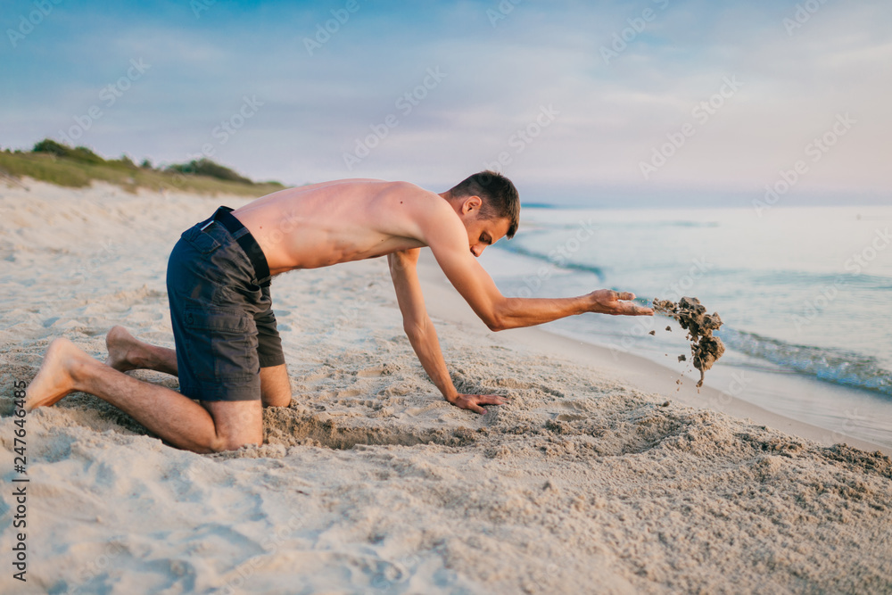 Young topless barefoot man in shorts digging hole by hands on beach beyond sea in summer evening. Funny boy playing on beach with sand on desolate cost near ocean ike child. Vacation leisure and hobby Young topless barefoot man in shorts digging hole by hands on beach beyond sea in summer evening. Funny boy playing on beach with sand on desolate cost near ocean ike child. Vacation leisure and hobby