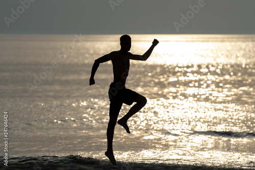 Dark silhoutte of man jumping with hands apart like superhero over sea with sun light on background. Unrecognizable man dancing beyond sea at sunset in summer evening. Freedom and happiness concept.
