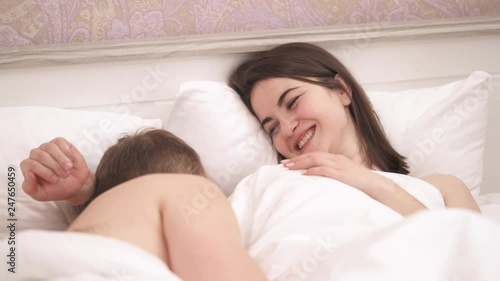 Loving couple lying on the bed under blanket. The girl offers a condom. Man opens it with his teeth