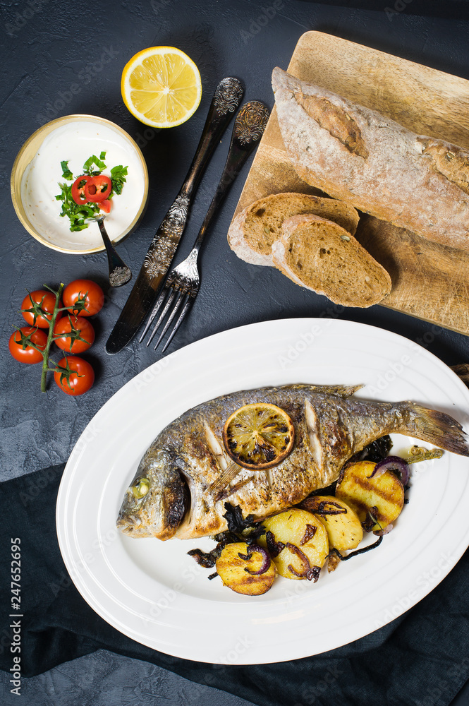 Baked Dorado fish on a white plate, garnish fried potatoes. Dark background, top view