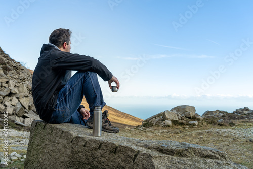 Middle age man sitting on the rock drinking a tea or coffee in cold morning looking on the vale and sea from mountain range, view from behind