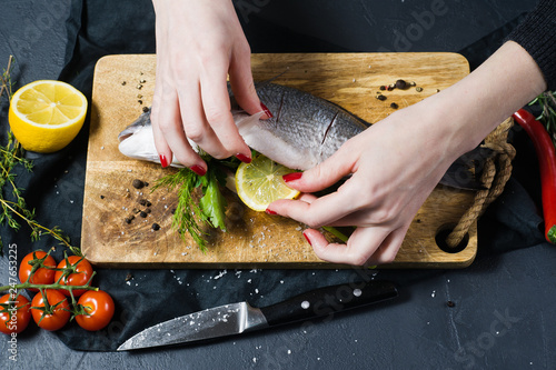 The chef's hands stuffed Dorado fish on a wooden chopping Board with parsley and cilantro. Ingredient rosemary, tomatoes, chili, lemon, thyme, pepper, salt. Dark background, side view
