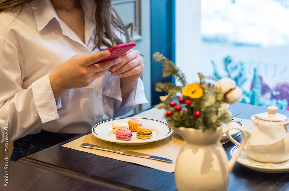 A woman with a smartphone is taking a picture of a macaroons and a cup of tea in a cafe. A woman with a smartphone is taking a picture of a macaroons and a cup of tea in a cafe.