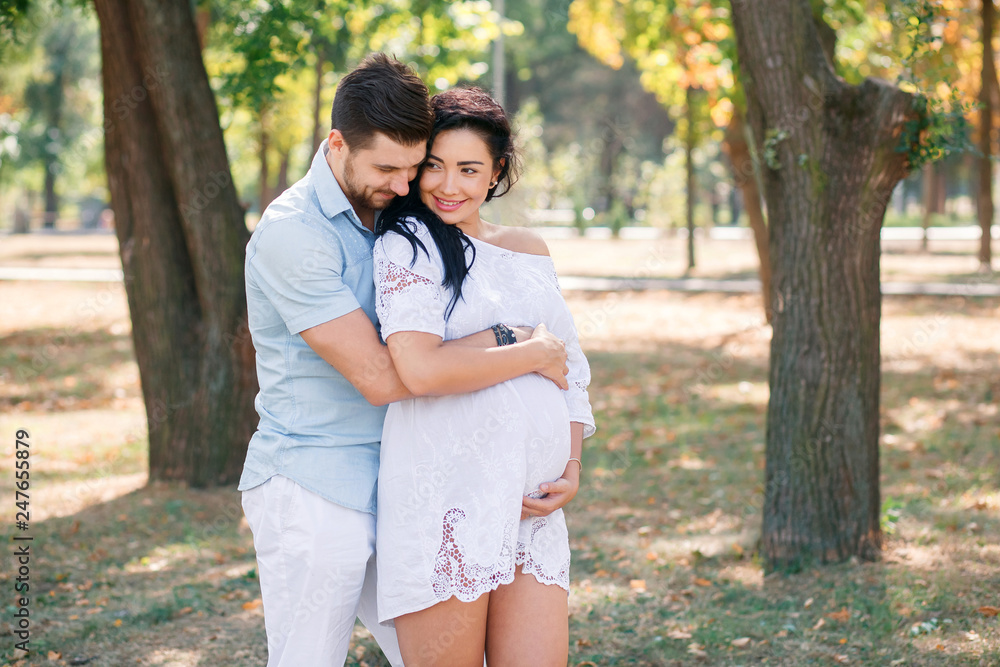 Pregnant girl walks in the park with her husband, hugging and kissing, enjoying the beautiful weather