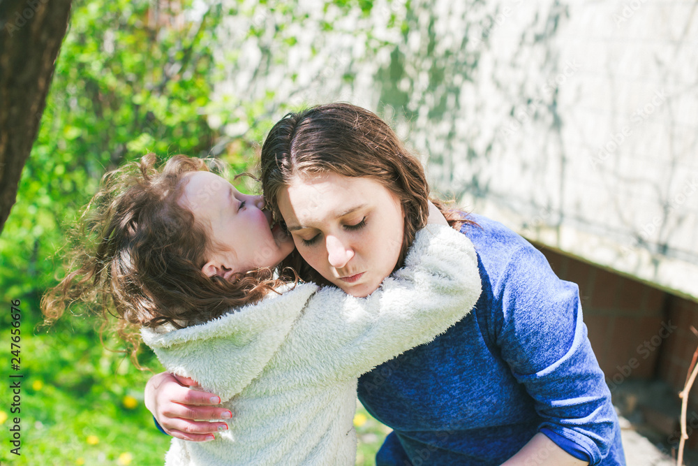 Fototapeta premium Little girl hugs mother sitting on the grass. Child, playing, threw his mother on grass. Family in spring walks on street.