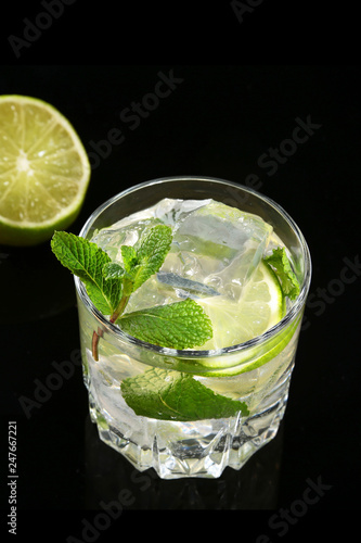 Glass of cocktail with rum, lime, ice cubes and mint leaves on black mirror background.