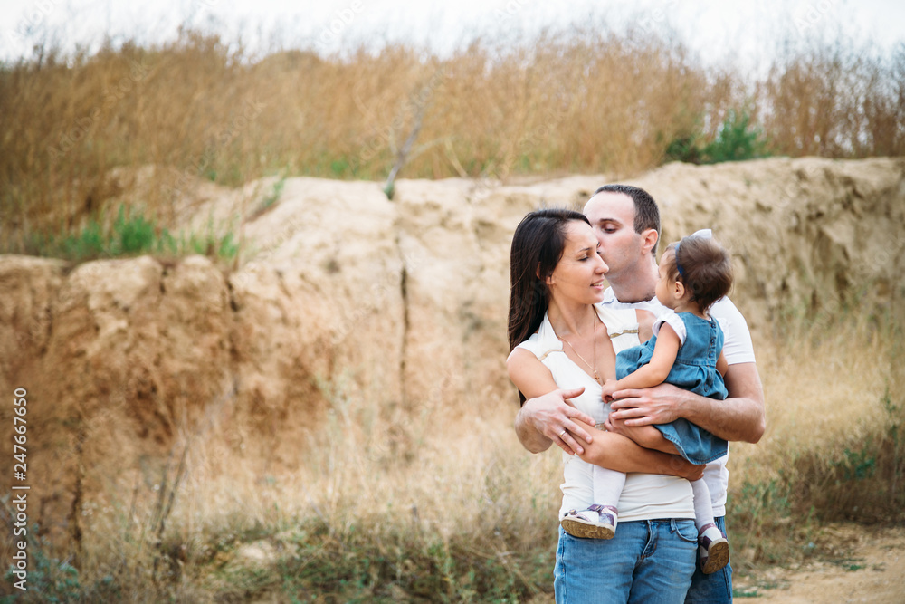 Happy young family in white t-shirts and blue jeans with a small daughter in dress hugging, outdoors background Happy young family in white t-shirts and blue jeans with a small daughter in dress hugging, outdoors background