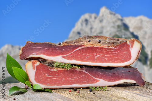 Südtiroler Speck auf einem Holztisch vor Bergen in den Alpen – Typical South Tyrolean bacon divided into two halfes lying on a rustic table in front of mountains of the alps photo
