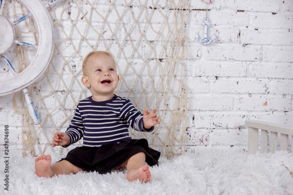Fototapeta premium Portrait of a cute and naughty european little girl in a sailors clothing in the studio of marine style on a background of white brick wall