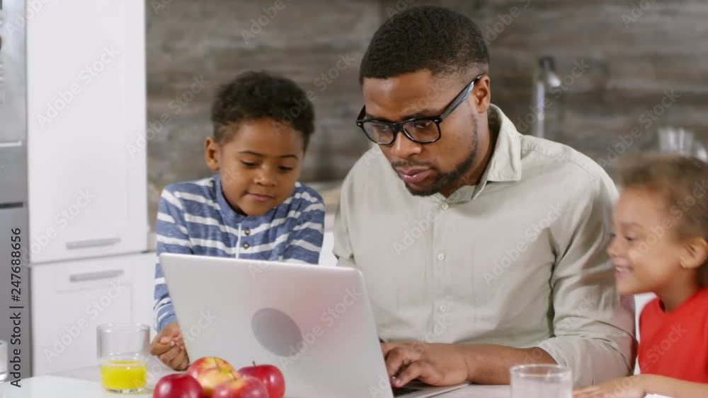 Tracking right of African man sitting at laptop in kitchen, typing while his kids coming to him and starting watching what he is doing