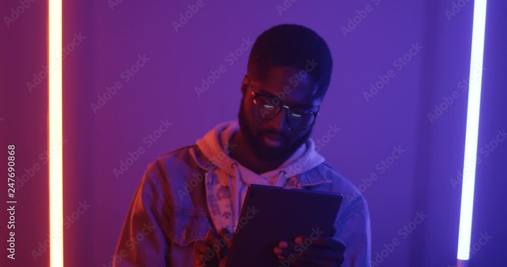 Young African American guy in glasses standing in front of the camera and using tablet device with smile on his face on the backgroung wall with neon lights. Young African American guy in glasses standing in front of the camera and using tablet device with smile on his face on the backgroung wall with neon lights.