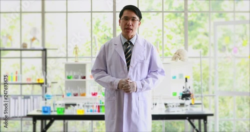 Asian scientist standing and talking to introducing his laboratory to the camera, colorful test tubes on table behind him.