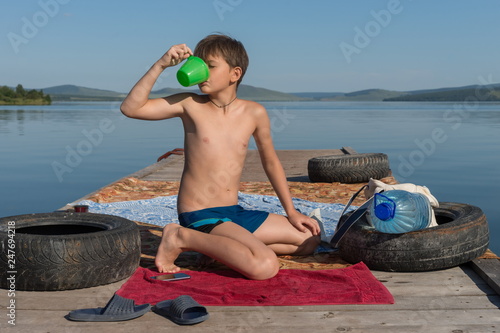 A boy of 11 years old slake his thirst with water from a mug, sitting on a wooden pier, against the backdrop of the  lake on a summer sunny day.