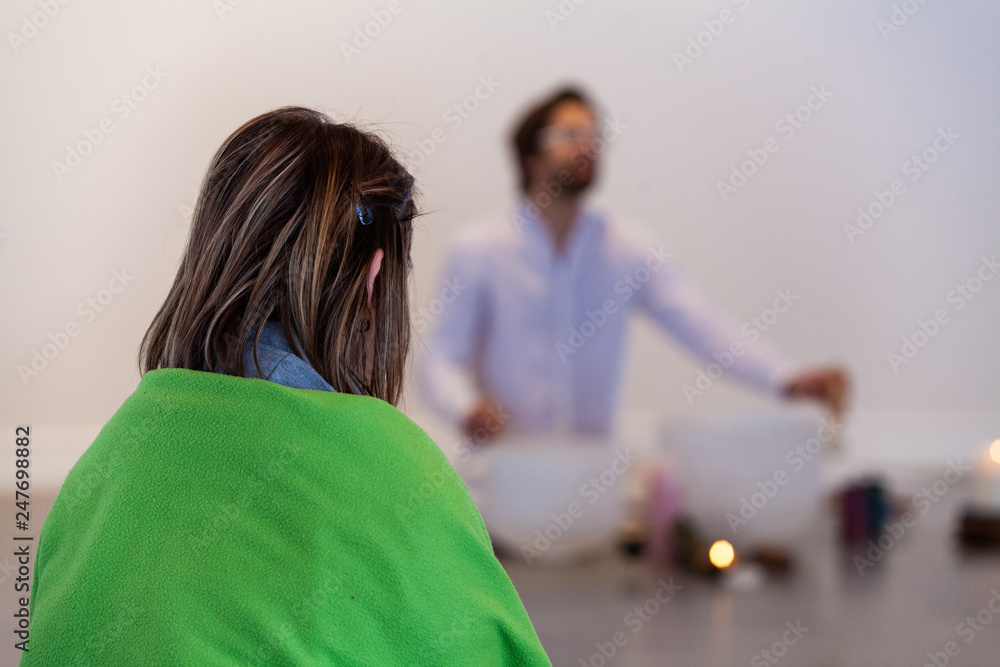 Woman listening to crystal bowls. Woman seen from behind, wrapped in a blanket, listening to a man dressed in white playing crystal bowls and singing in the blurry background