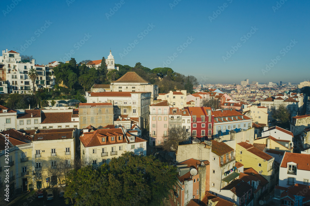 Obraz premium Beautiful panoramic Lisbon landmark with tiny houses with moorish architectural elements on the hill; drone view of picturesque labyrinth of narrow streets and small squares in early morning, Portugal