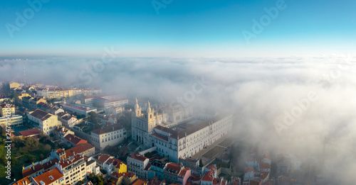 Aerial  misty morning in ancient Alfama district of the Portuguese capital  drone view of picturesque labyrinth of narrow streets  sun rising up over clouds  unusual weather in touristic area  Lisboa