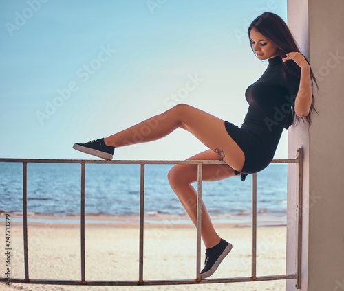 Young beautiful girl with a tattoo on her leg sitting on the railing against the beautiful sea coast