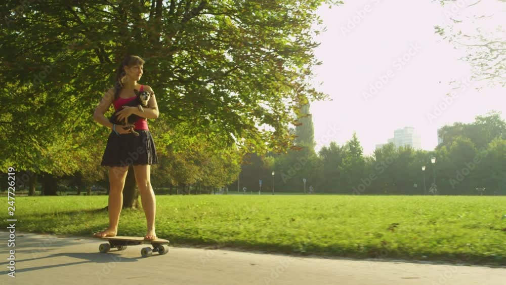SLOW MOTION, SUN FLARE: Cheerful young woman skating through the park while holding her cute puppy. Golden summer sunbeams shine on the happy Caucasian girl riding her high tech electric skateboard. SLOW MOTION, SUN FLARE: Cheerful young woman skating through the park while holding her cute puppy. Golden summer sunbeams shine on the happy Caucasian girl riding her high tech electric skateboard.