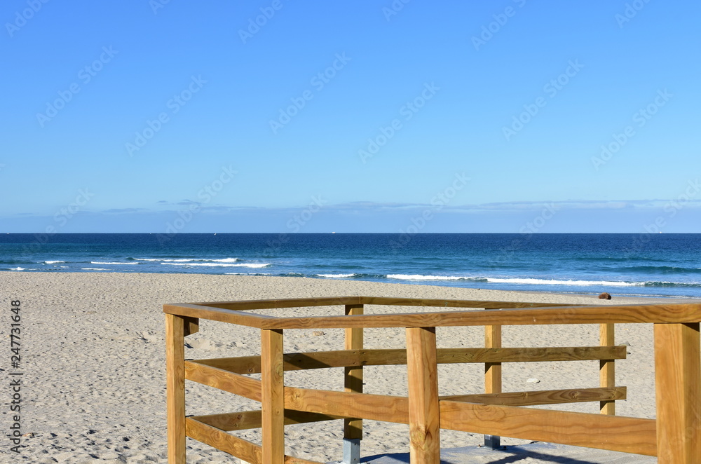 Beach with wooden handrail and morning light. Golden sand and blue sea with waves and white foam. Sunny day, Galicia, Spain.