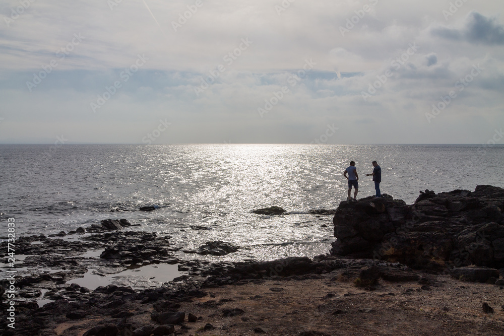 Silhouette of two men standing on a large rock by the ocean.