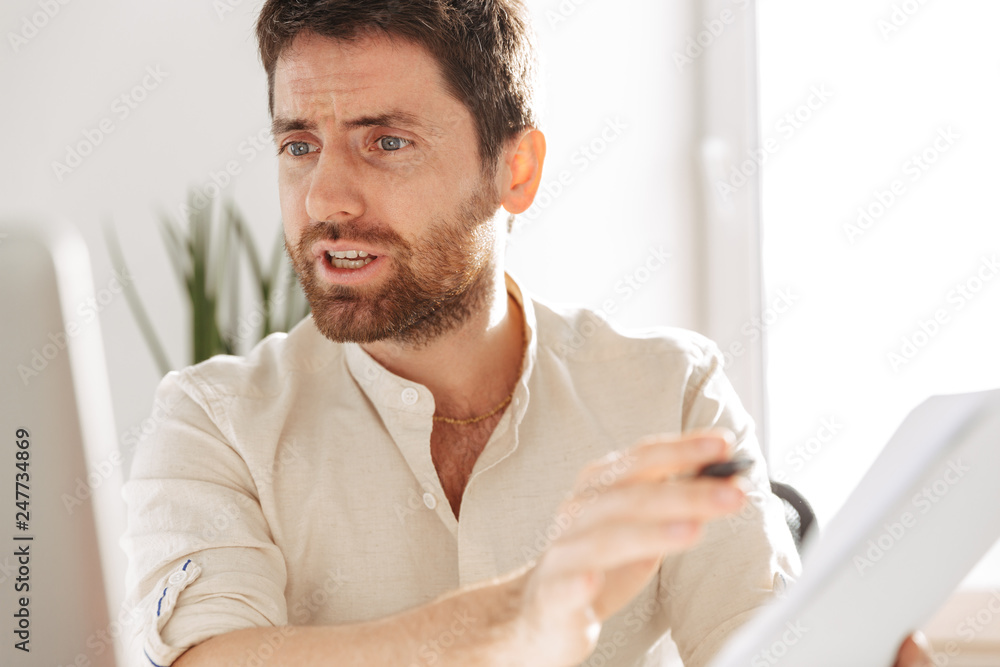 Photo of emotional office worker 30s wearing white shirt using laptop and paper documents, while sitting at table in modern workplace