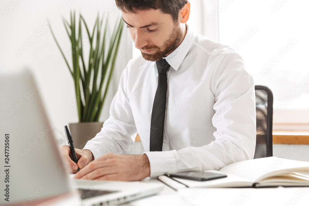 Fototapeta premium Image of successful office worker 30s wearing white shirt working with laptop and notebook, in modern workplace
